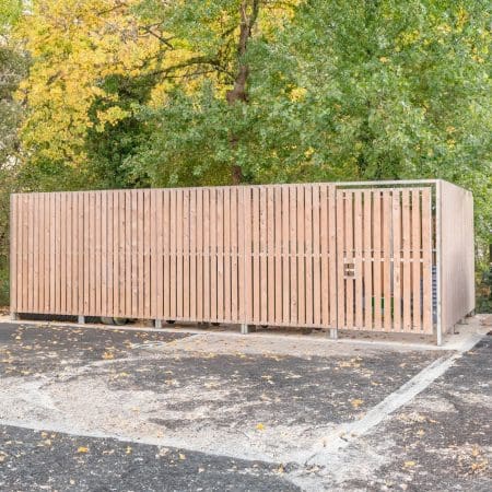 A wooden thurrock bin store enclosure is situated in a parking lot, with surrounding trees featuring green and yellow foliage. The enclosure has a gate and is designed to house waste and recycling bins. The ground is covered with asphalt, and some scattered leaves are visible.
