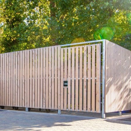 A large, wooden Thurrock Bin Store with vertical slats surrounds a structure. The store is set against a backdrop of green trees with sunlight filtering through the leaves. There is a small, dark-colored mailbox attached near the gate on the fence.