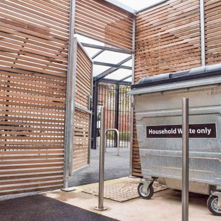 A large, gray trash bin labeled "Household Waste only" stands in a designated enclosure made of horizontal wooden slats. The area is partially covered with a metal frame and has an open gate leading out onto a paved pathway.