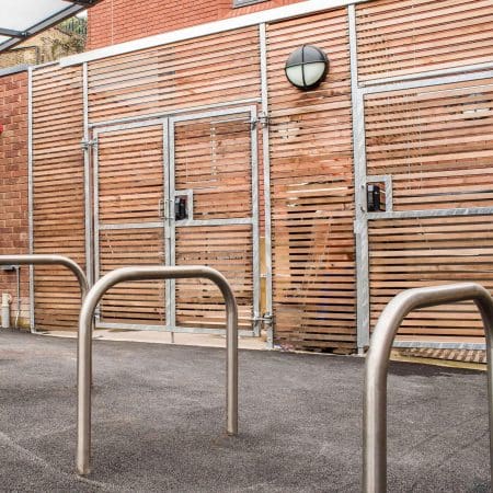 An outdoor area featuring a metal bike rack with three sheffield stands on a paved surface. Behind the bike rack, there is a wooden fence with horizontal slats and a metal frame, along with a circular wall-mounted light fixture above a door.