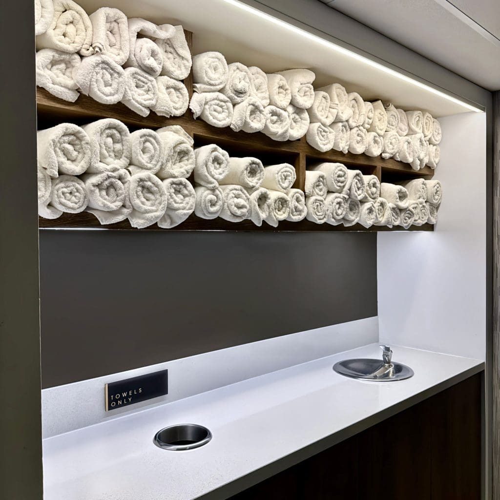 Close up of towel station with several towels rolled up and stored in the shelf above the counter. There is a sink and dirty towel slot installed and exposed wood panelling.